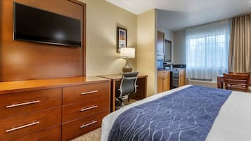 A hotel room with a large bed, a desk with a chair, a flat-screen TV, a lamp, and a view of the kitchen and bathroom through the window.