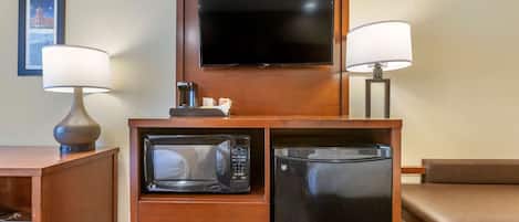A compact hotel room with a flat-screen TV mounted on a wooden cabinet, a microwave, a refrigerator, a wooden desk with a lamp, and a brown leather chair.