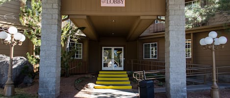 Una entrada de edificio con un letrero que dice “Lobby” sobre la puerta, un juego de escaleras que sube a la entrada, y un bote de basura al frente.