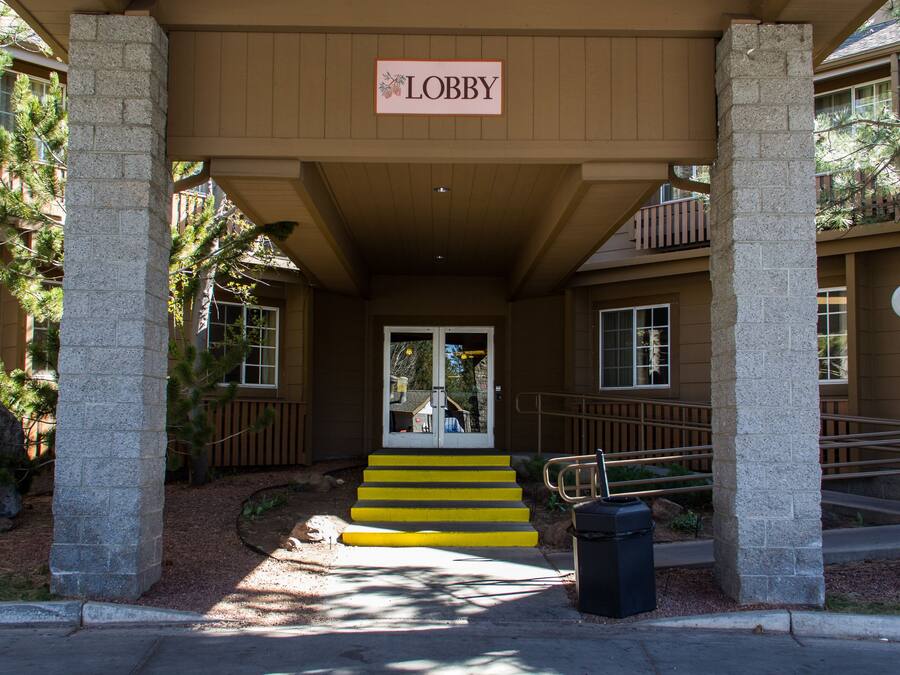 Una entrada de edificio con un letrero que dice “Lobby” sobre la puerta, un juego de escaleras que sube a la entrada, y un bote de basura al frente.
