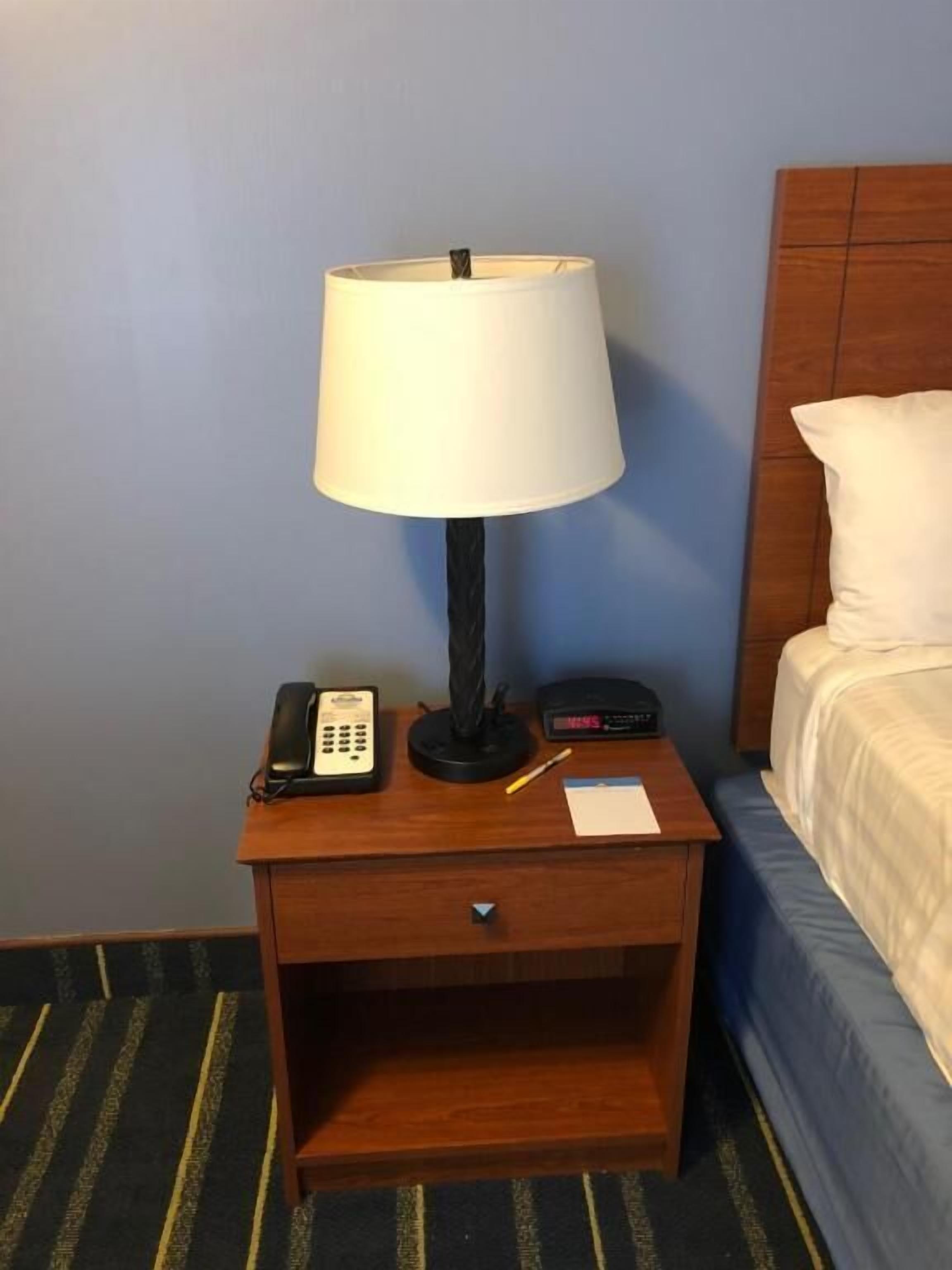 Une table de chevet en bois, avec une lampe blanche, un téléphone et une horloge numérique.