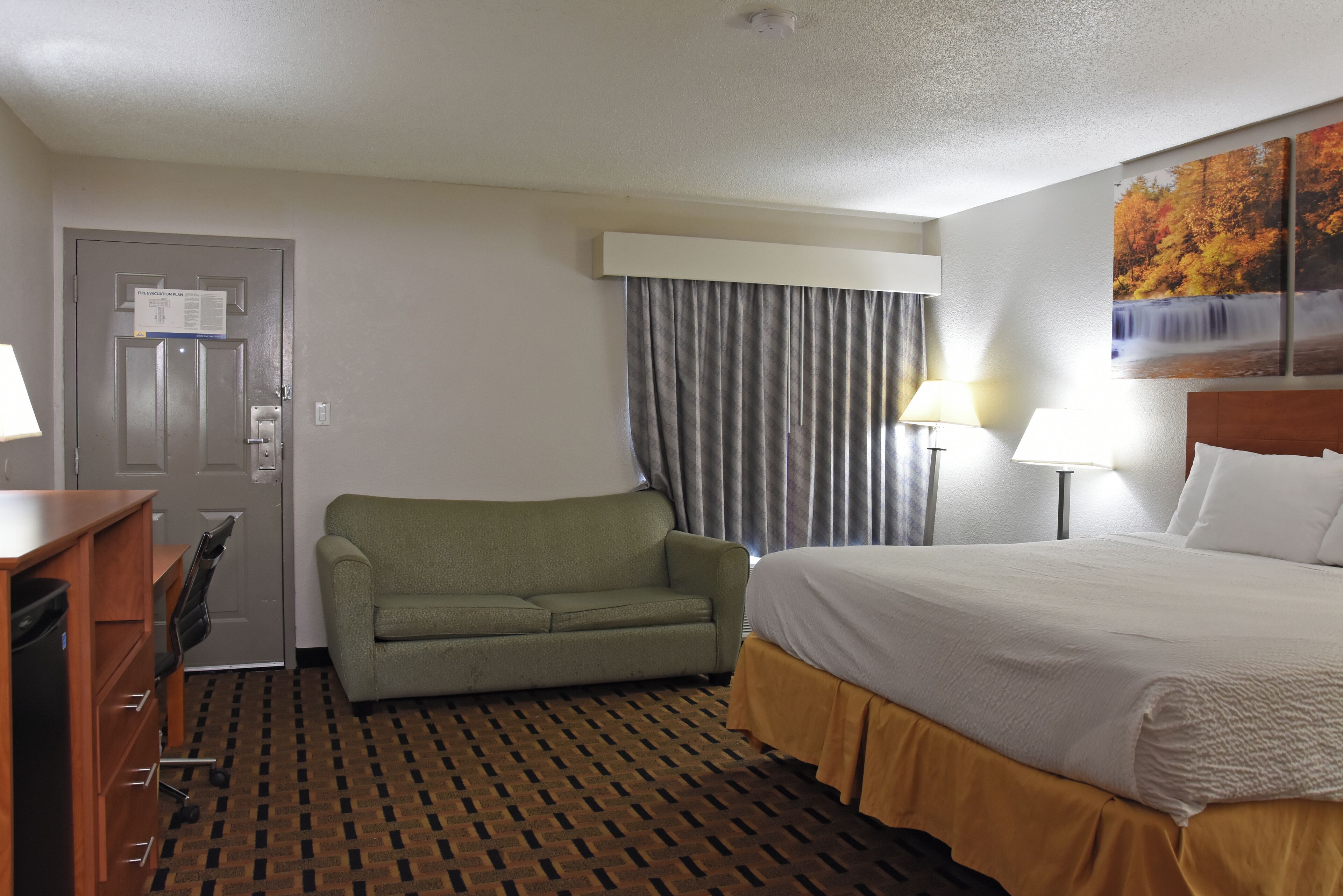 A hotel room with a bed, a sofa, a desk with a chair, a lamp, and a painting on the wall.