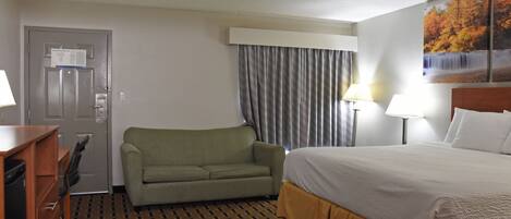 A hotel room with a bed, a sofa, a desk with a chair, a lamp, and a painting on the wall.