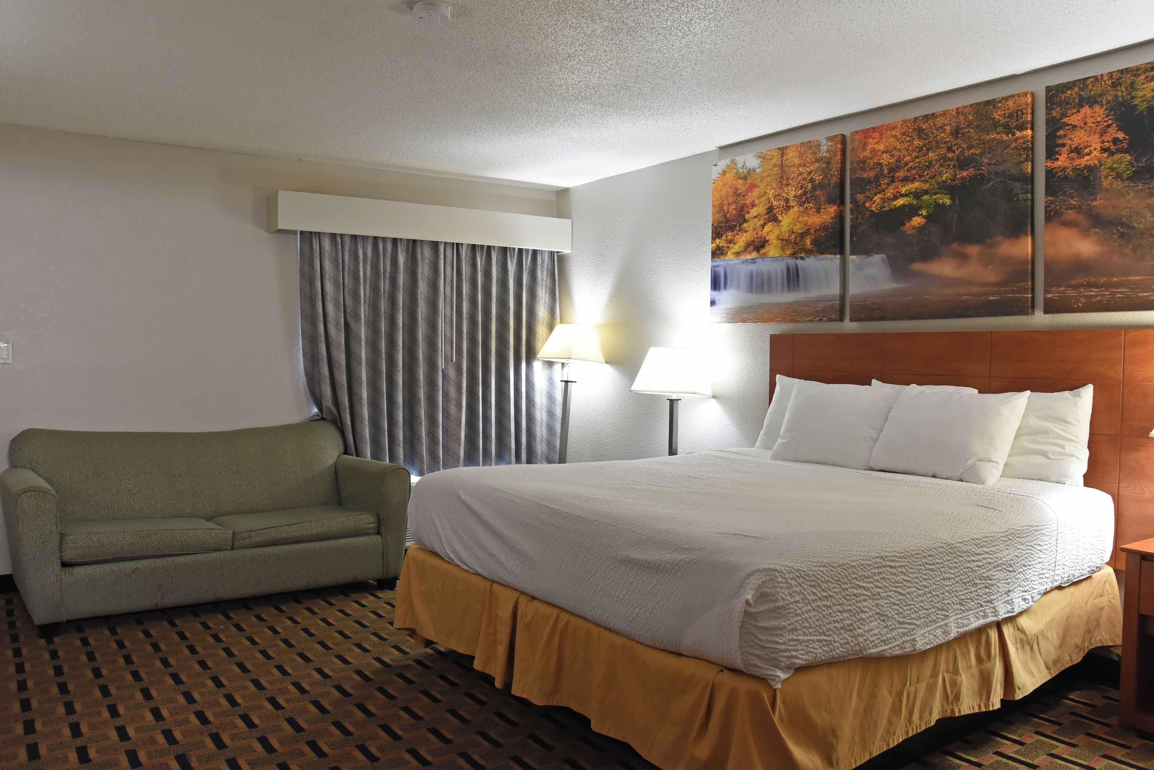 A hotel room with a bed, a sofa, a painting of a waterfall, and a patterned carpet.