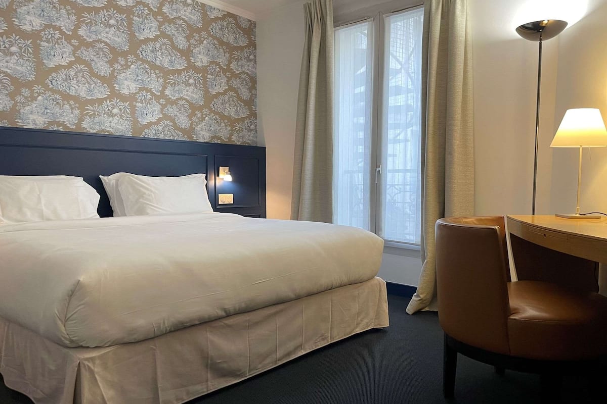 Habitación de hotel con una cama grande, un escritorio con silla, una lámpara y una ventana con cortinas.
