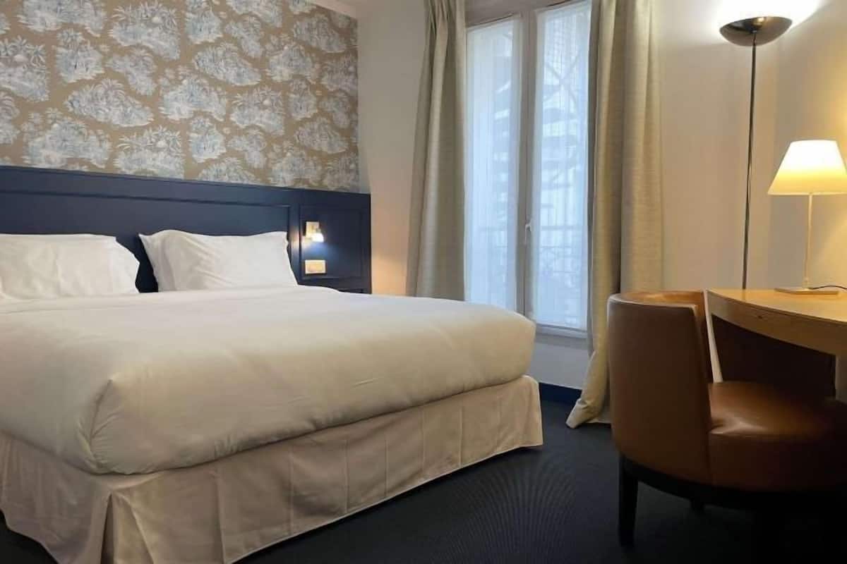 Una habitación de hotel con cama, escritorio, silla, lámpara y ventana con cortinas.