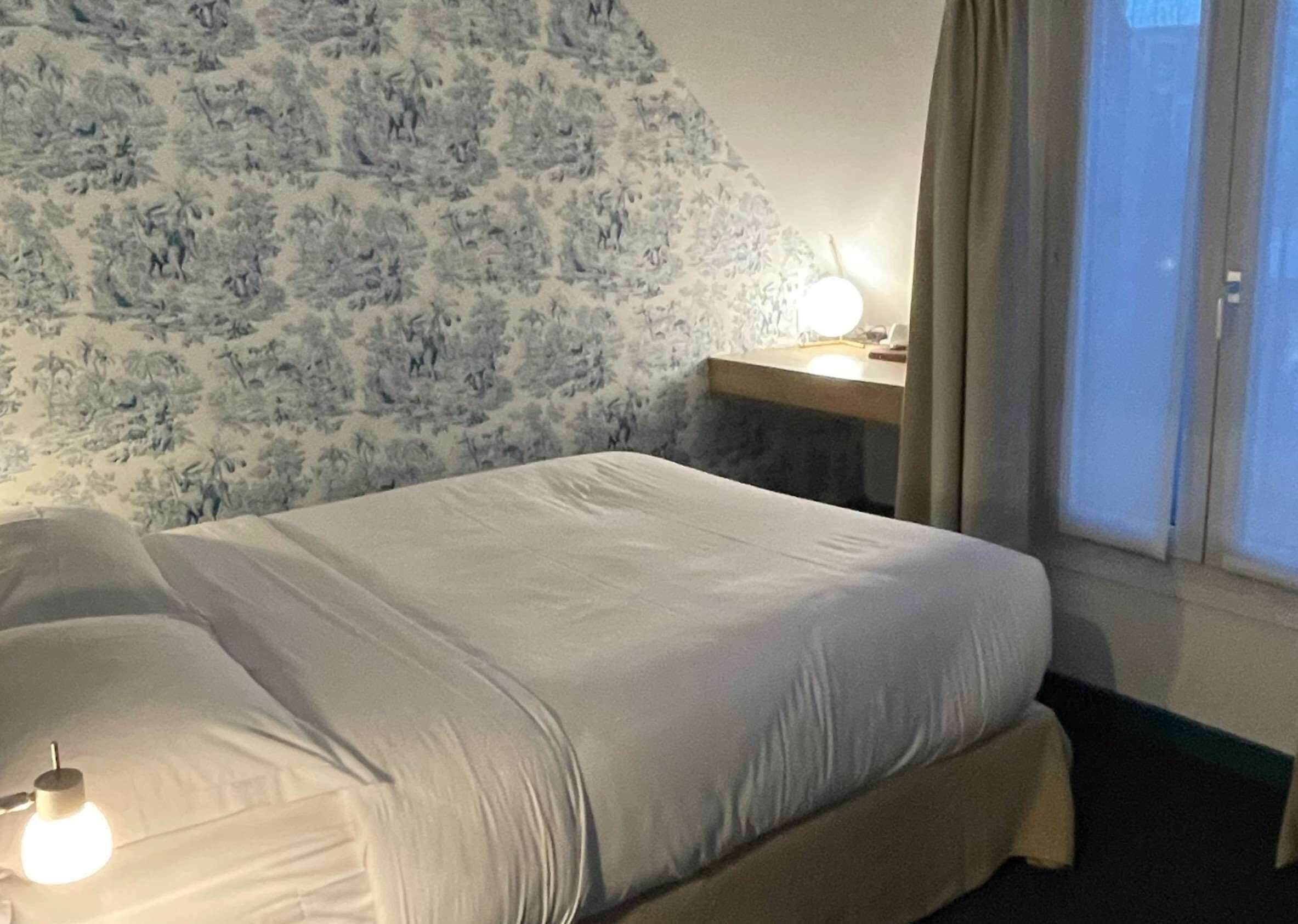 Habitación de hotel con cama, mesita de noche con lámpara, ventana con cortinas y pared con estampado floral.