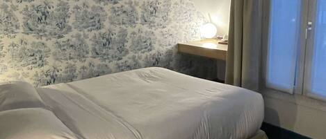 Habitación de hotel con cama, mesita de noche con lámpara, ventana con cortinas y pared con estampado floral.