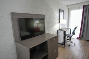 Habitación de hotel con televisión de pantalla plana montada en un mueble de madera, escritorio con silla y un ventanal con cortinas.