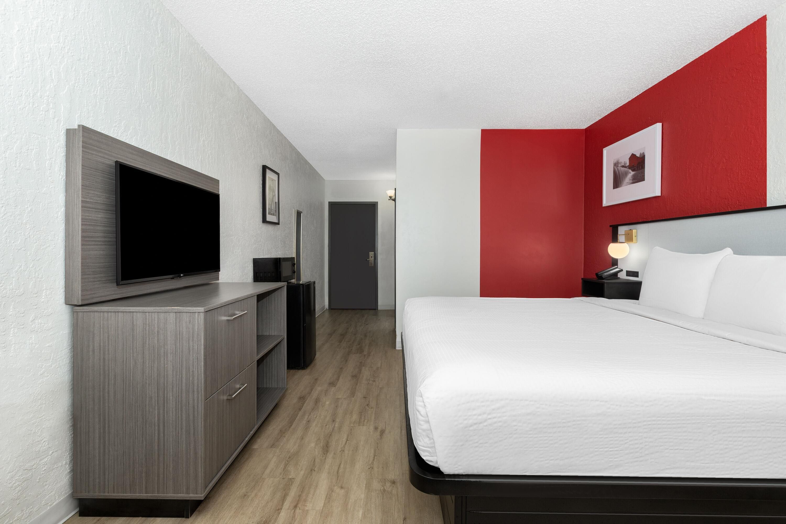 Quarto de hotel com cama, televisão, escrivaninha e uma parede em tom de vermelho.