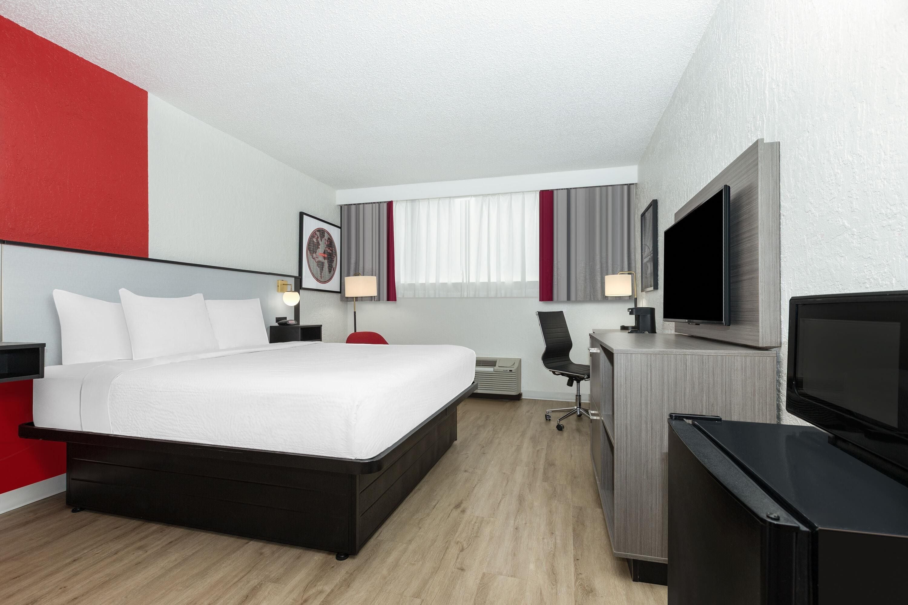A modern hotel room with a large bed, a desk with a chair, a flat-screen TV, and a large window with curtains.