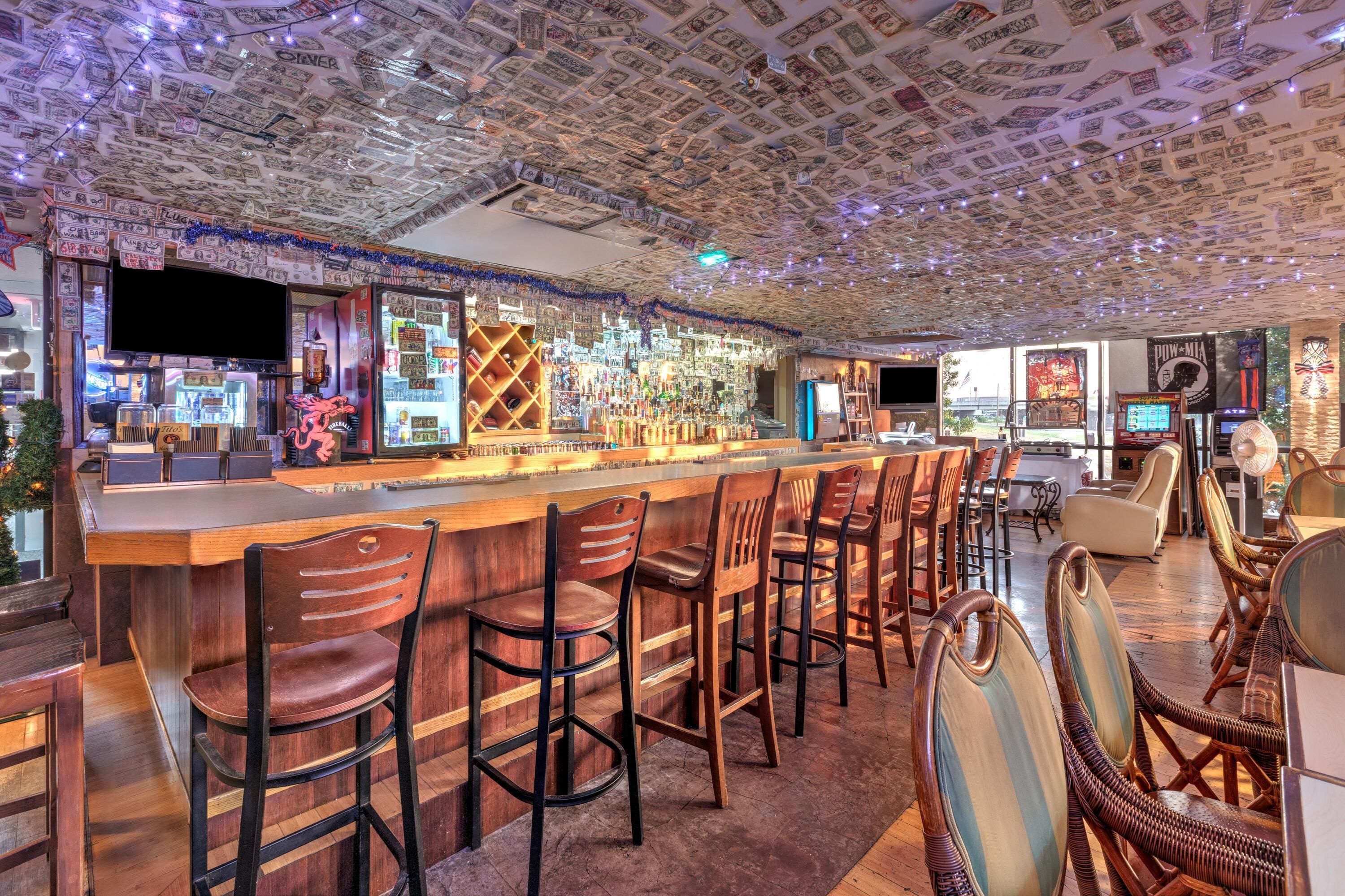 Un bar con techo cubierto de billetes de dólar, taburetes de madera y una variedad de botellas y vasos.