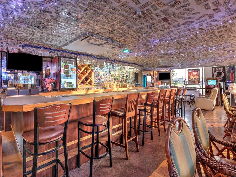 A bar with a ceiling covered in dollar bills, wooden bar stools, and a variety of bottles and glasses.