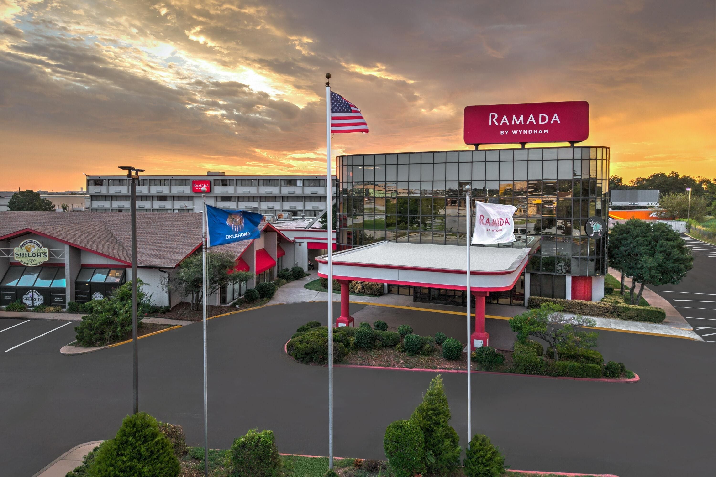 Un edificio comercial con un amplio frente de vidrio, un toldo rojo y blanco, y varias banderas, incluyendo la bandera estadounidense y un banderín de Ramada by Wyndham.