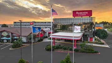 Un edificio comercial con un amplio frente de vidrio, un toldo rojo y blanco, y varias banderas, incluyendo la bandera estadounidense y un banderín de Ramada by Wyndham.