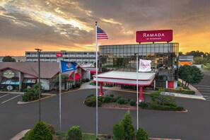 A commercial building with a large glass facade, a red and white awning, and multiple flags including the American flag and a banner for Ramada by Wyndham. - Ramada by Wyndham Tulsa Broken Arrow (Broken Arrow)