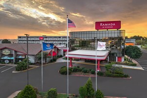 Un edificio comercial con un amplio frente de vidrio, un toldo rojo y blanco, y varias banderas, incluyendo la bandera estadounidense y un banderín de Ramada by Wyndham.