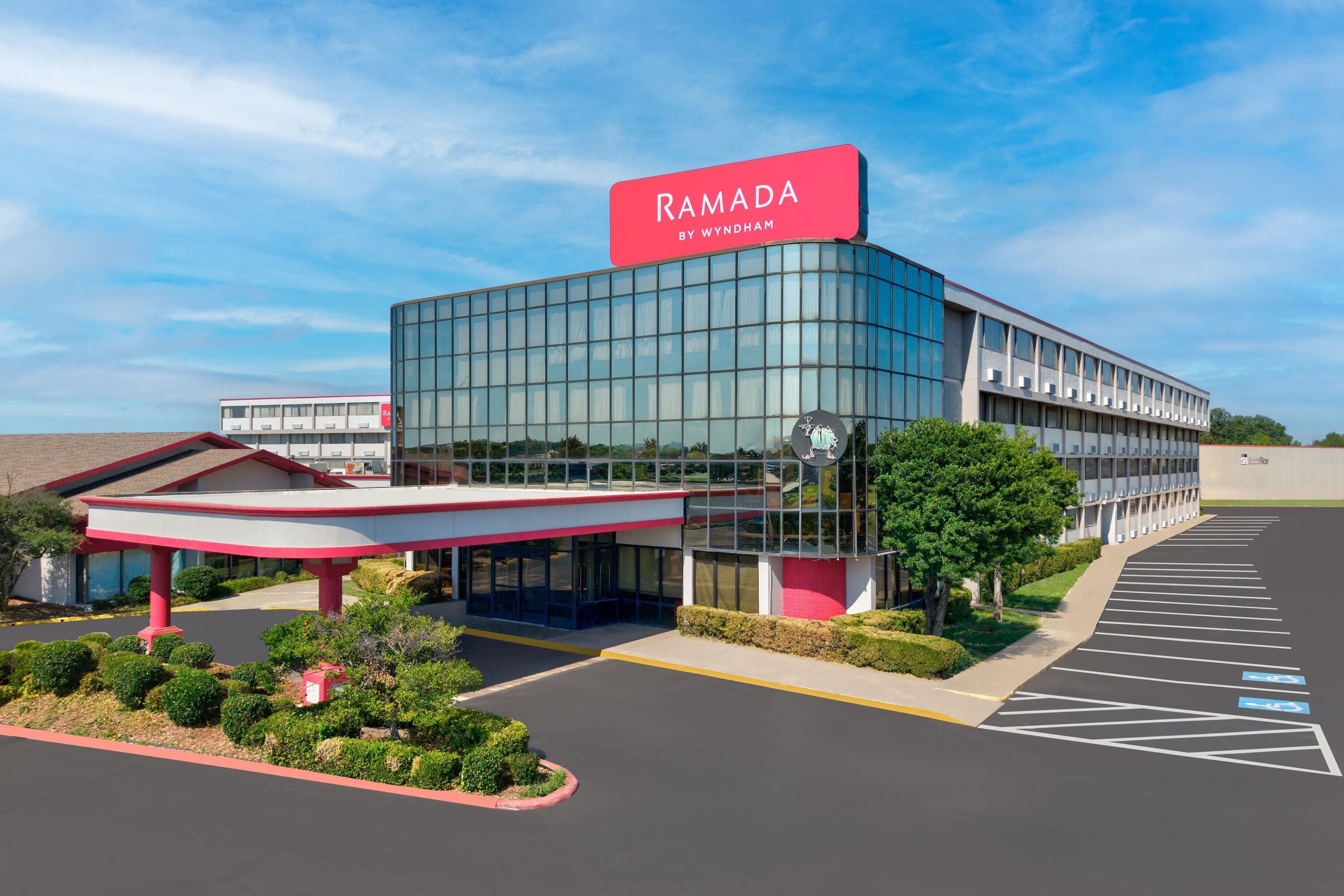 A modern building with a glass facade, a prominent red sign reading 'RAMADA BY WYNDHAM', and a parking lot with marked spaces.