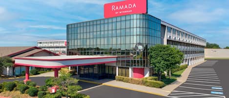 A modern building with a glass facade, a prominent red sign reading 'RAMADA BY WYNDHAM', and a parking lot with marked spaces.