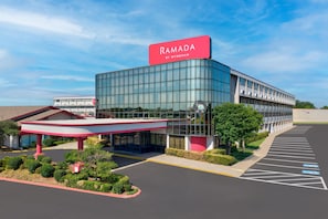 A modern building with a glass facade, a prominent red sign reading 'RAMADA BY WYNDHAM', and a parking lot with marked spaces. - Ramada by Wyndham Tulsa Broken Arrow (Broken Arrow)
