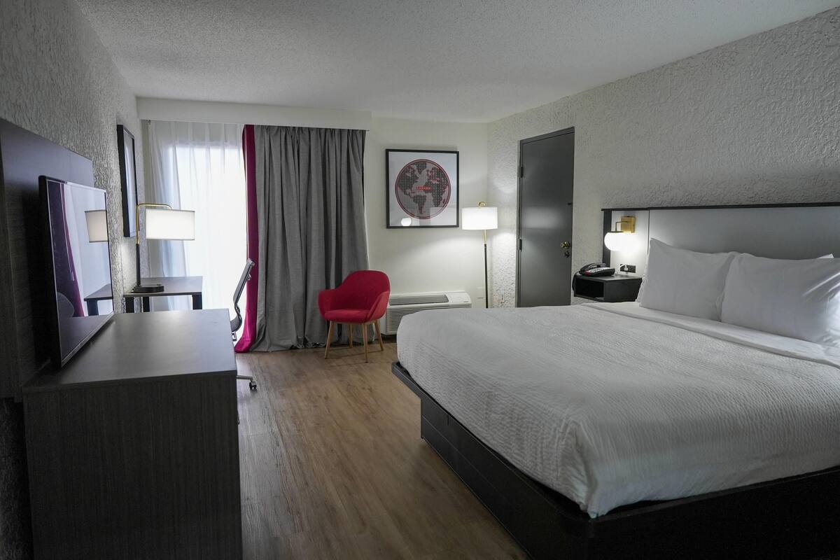 Una habitación de hotel con una cama, un escritorio, una silla, una lámpara, una obra de arte con un globo terráqueo y una puerta.