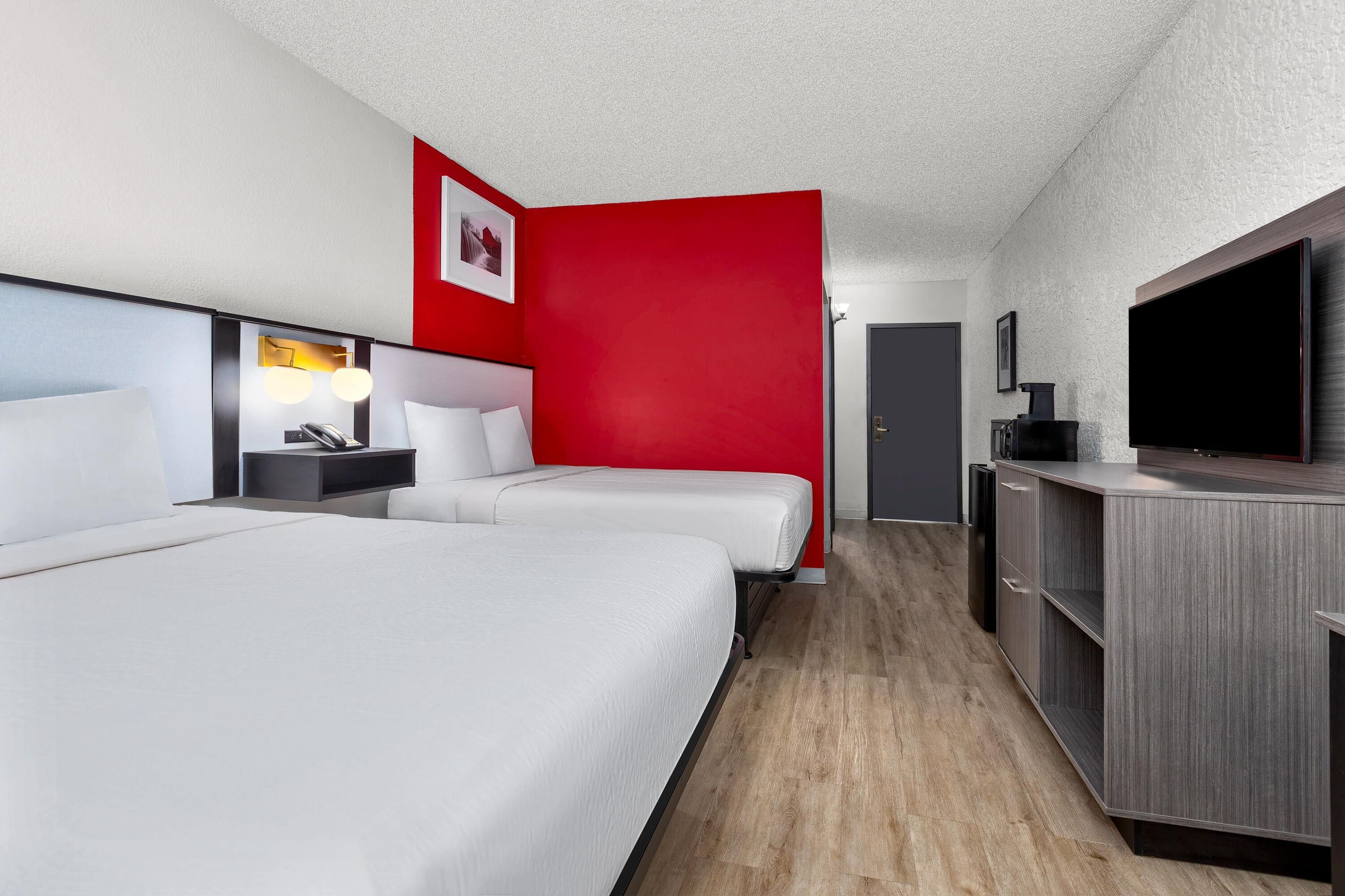 A hotel room with two beds, a desk with a lamp, a TV, and a red accent wall.