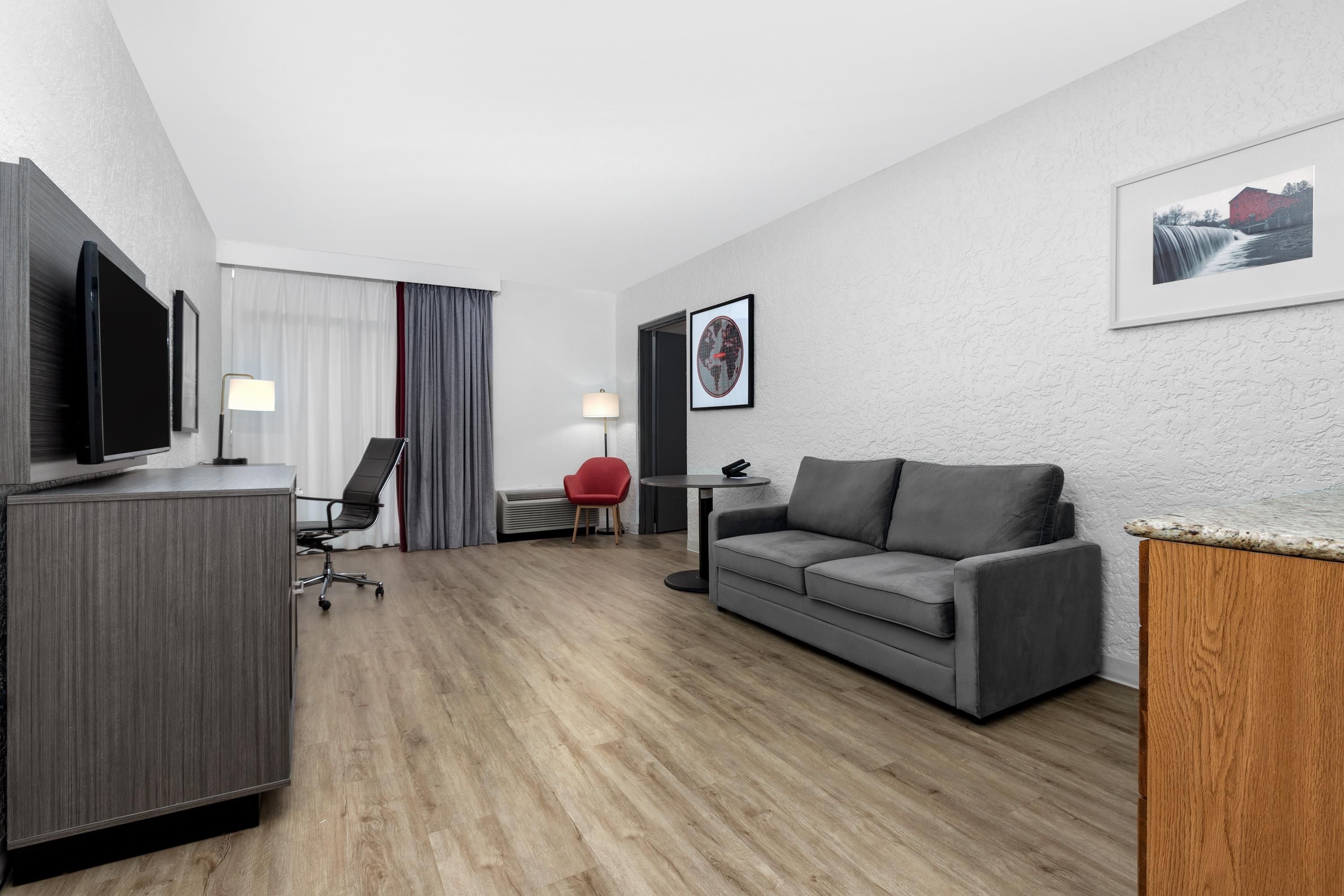 Quarto de hotel com televisão de tela plana, sofá cinza, cadeira vermelha, mesa pequena e quadro na parede.