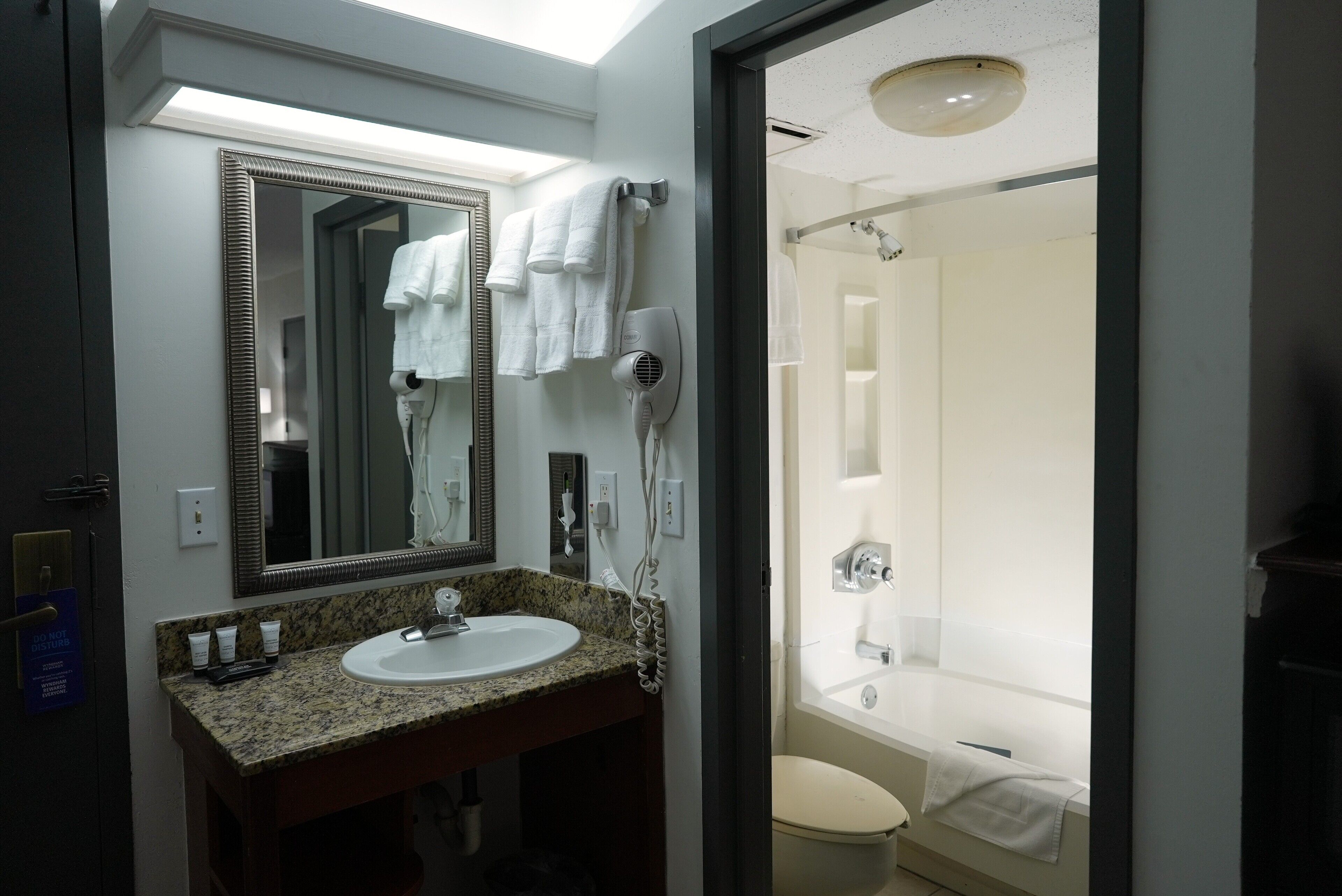 A bathroom with a granite countertop sink, a large mirror, a hairdryer, and a shower with a white towel.