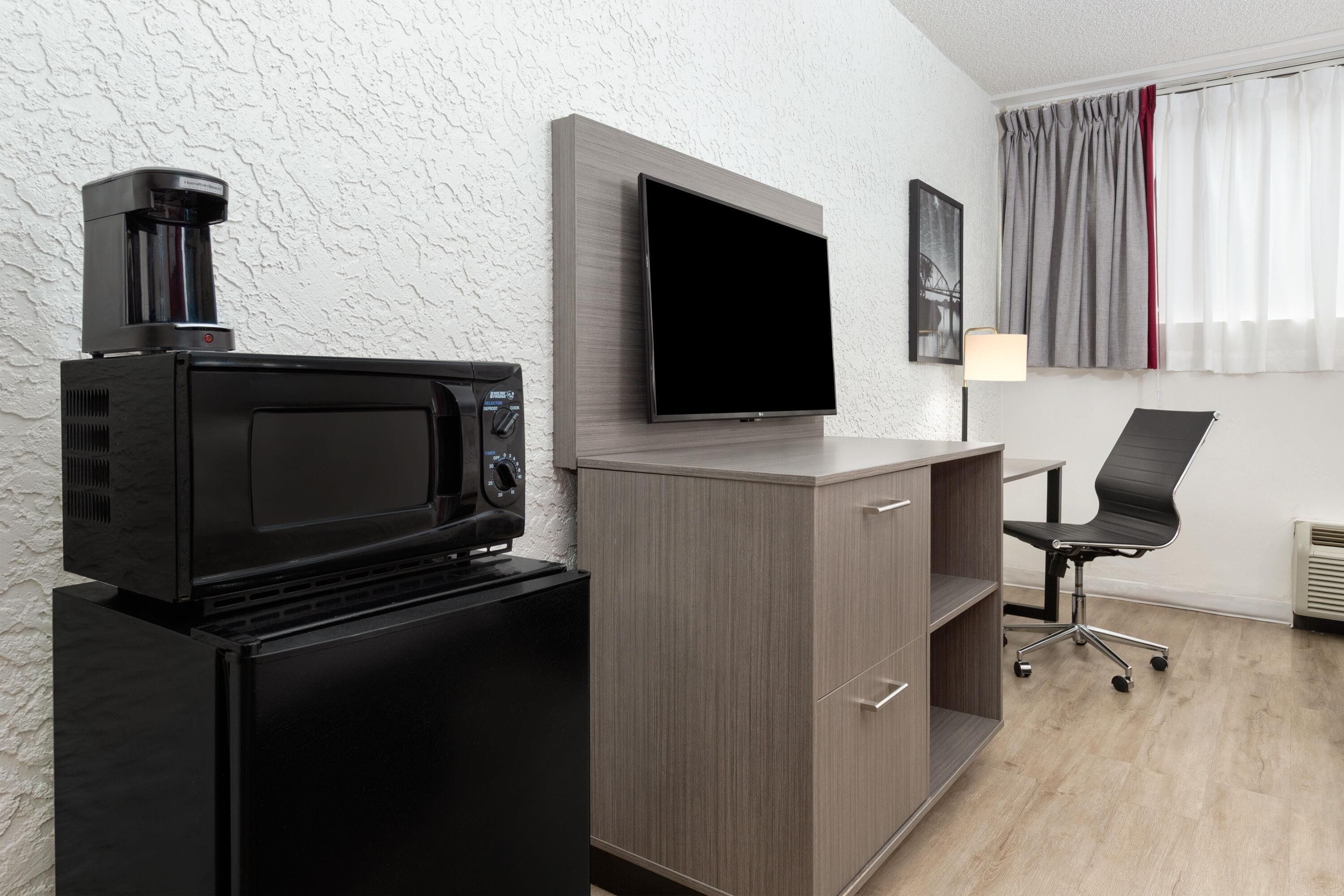 Una habitación compacta con kitchenette que incluye un microondas, un refrigerador pequeño y un televisor de pantalla plana montado en la pared. Cuenta con un escritorio con silla y lámpara, además de un ventanal con cortinas.