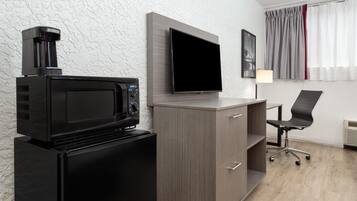 Una habitación compacta con kitchenette que incluye un microondas, un refrigerador pequeño y un televisor de pantalla plana montado en la pared. Cuenta con un escritorio con silla y lámpara, además de un ventanal con cortinas.