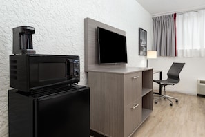 A compact room with a kitchenette area featuring a microwave, a small refrigerator, and a flat-screen TV mounted on the wall. There is a desk with a chair and a lamp, and a window with curtains.