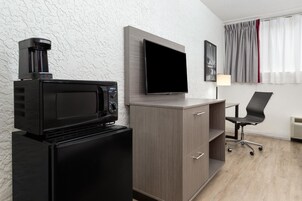 Una habitación compacta con kitchenette que incluye un microondas, un refrigerador pequeño y un televisor de pantalla plana montado en la pared. Cuenta con un escritorio con silla y lámpara, además de un ventanal con cortinas.