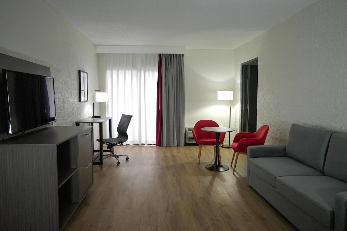 Una habitación de hotel con una televisión de pantalla plana, un sofá gris, una silla roja, una mesita y un escritorio con silla.