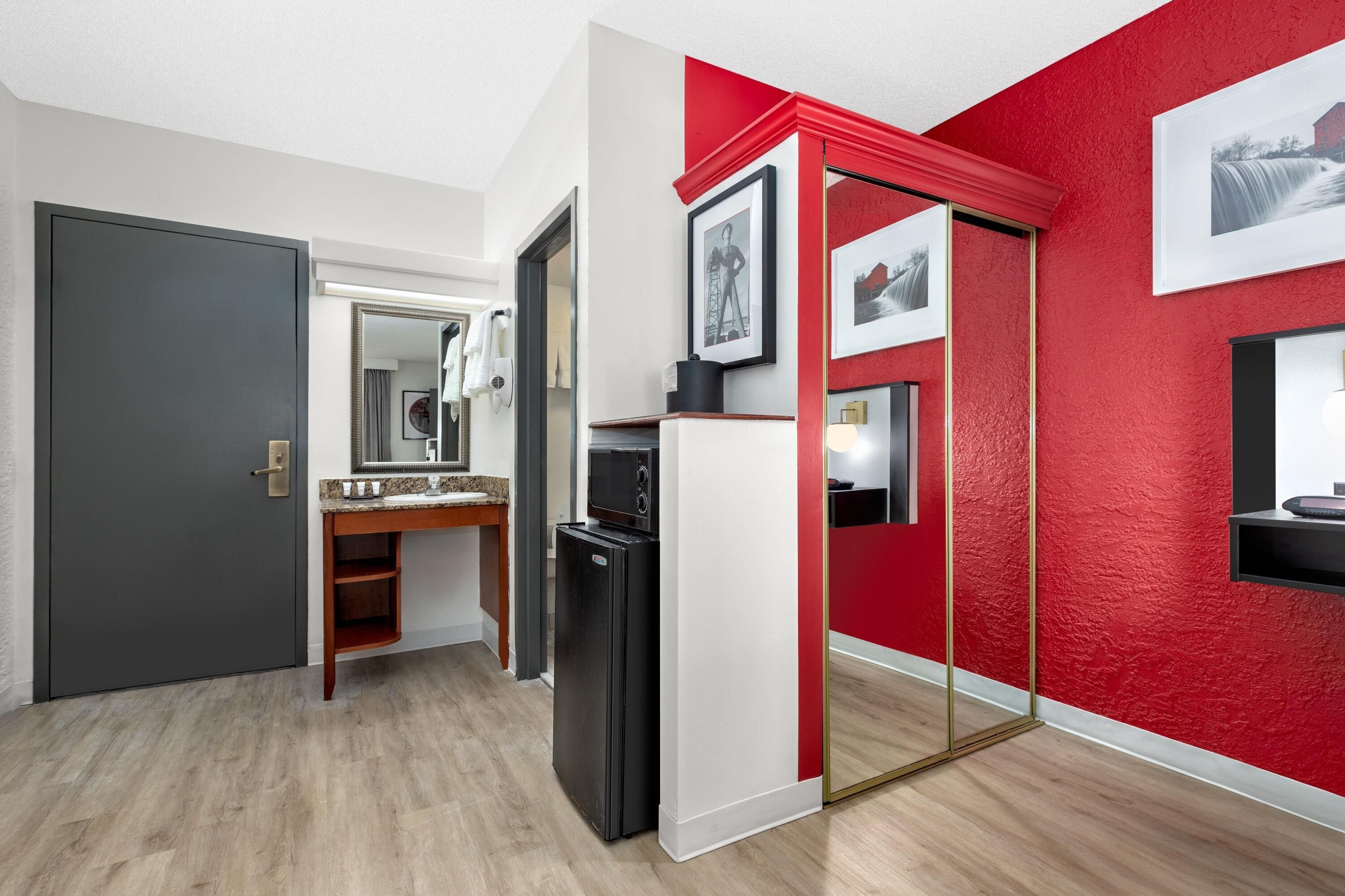 Habitación de hotel moderna con puerta gris, tocador de madera con espejo, refrigerador negro y una pared de acento roja.