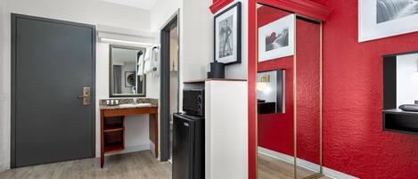 A modern hotel room with a grey door, a wooden vanity with a mirror, a black refrigerator, and a red accent wall.