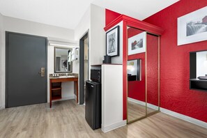 A modern hotel room with a grey door, a wooden vanity with a mirror, a black refrigerator, and a red accent wall. - Ramada by Wyndham Tulsa Broken Arrow (Broken Arrow)