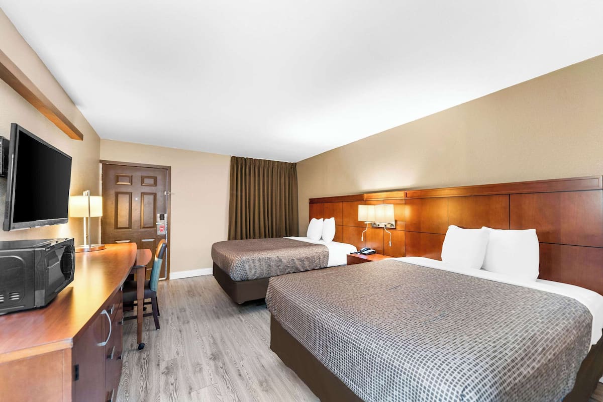 A hotel room with two beds, a desk with a flat-screen TV, a minibar, and a door with a curtain.