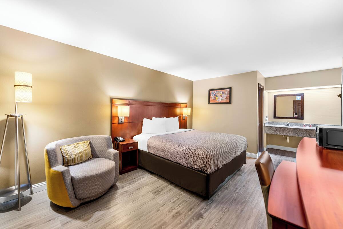 A hotel room with a bed, a chair, a desk, a nightstand, a lamp, and a bathroom mirror.