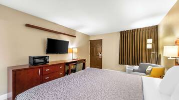 A hotel room with a bed, a desk with a television, a chair, and a lamp.