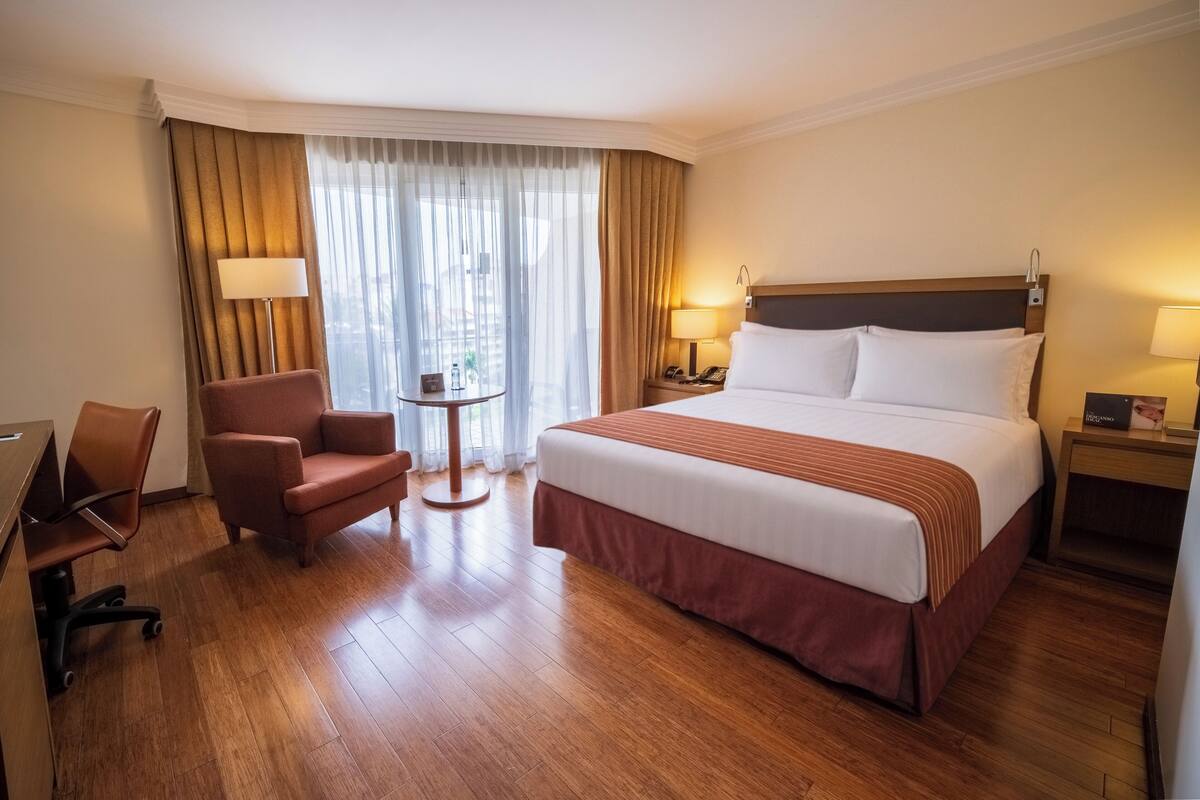 A hotel room with a large bed, a desk, a chair, a small table, and a nightstand with a lamp.