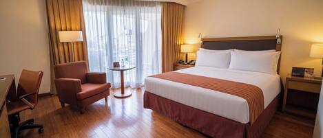 A hotel room with a large bed, a desk, a chair, a small table, and a nightstand with a lamp.