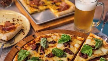 Une pizza garnie de fromage, de basilic et de tomates séchées au soleil, présentée sur une planche en bois, un pichet de bière et une autre pizza sur un plateau.