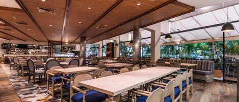 Un restaurant spacieux avec des tables en bois et des chaises rembourrées bleues, de grandes fenêtres offrant une vue sur la verdure et un design moderne au niveau du plafond.