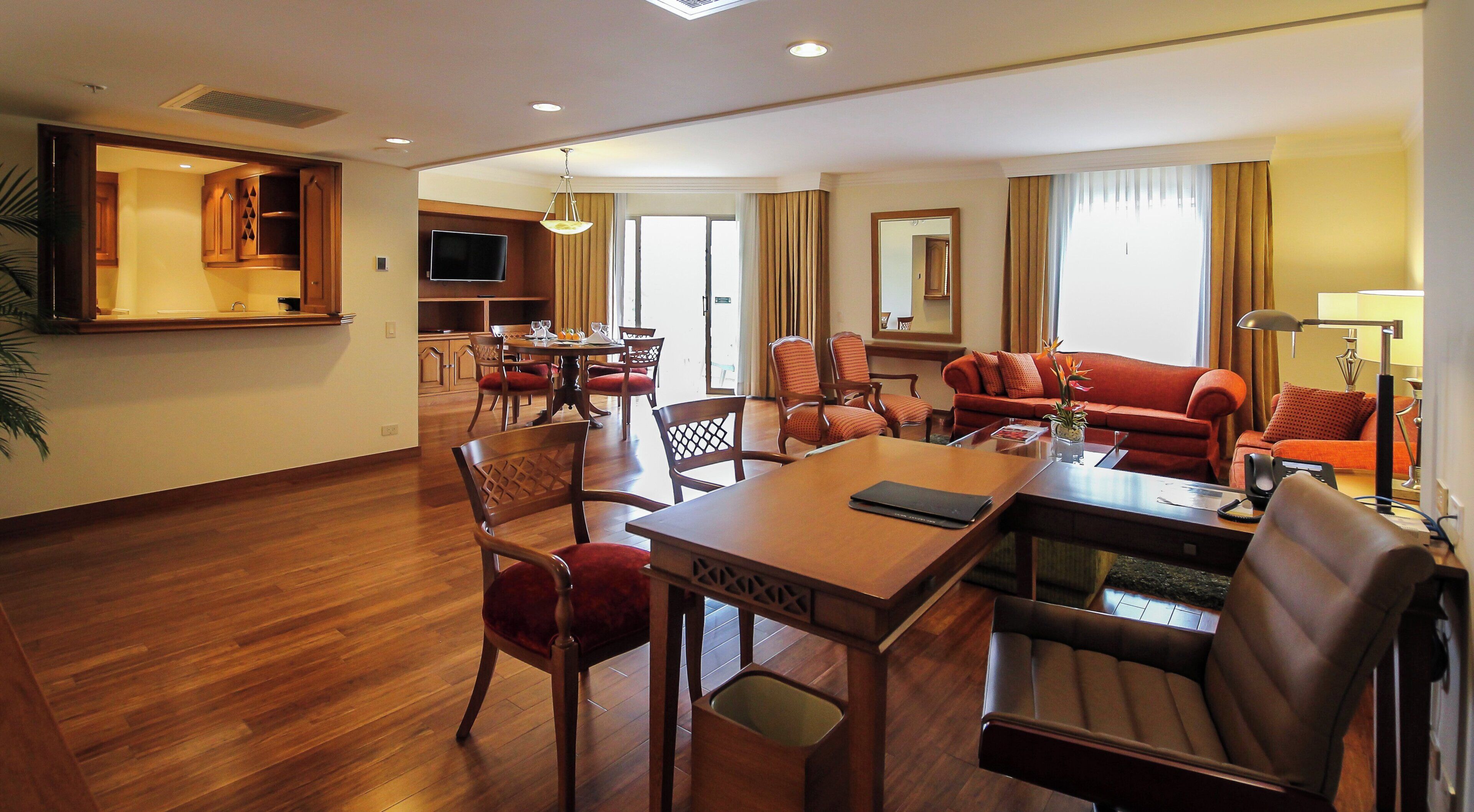 A hotel room with a dining area, a living room with a sofa and armchairs, a TV, and a kitchen area.