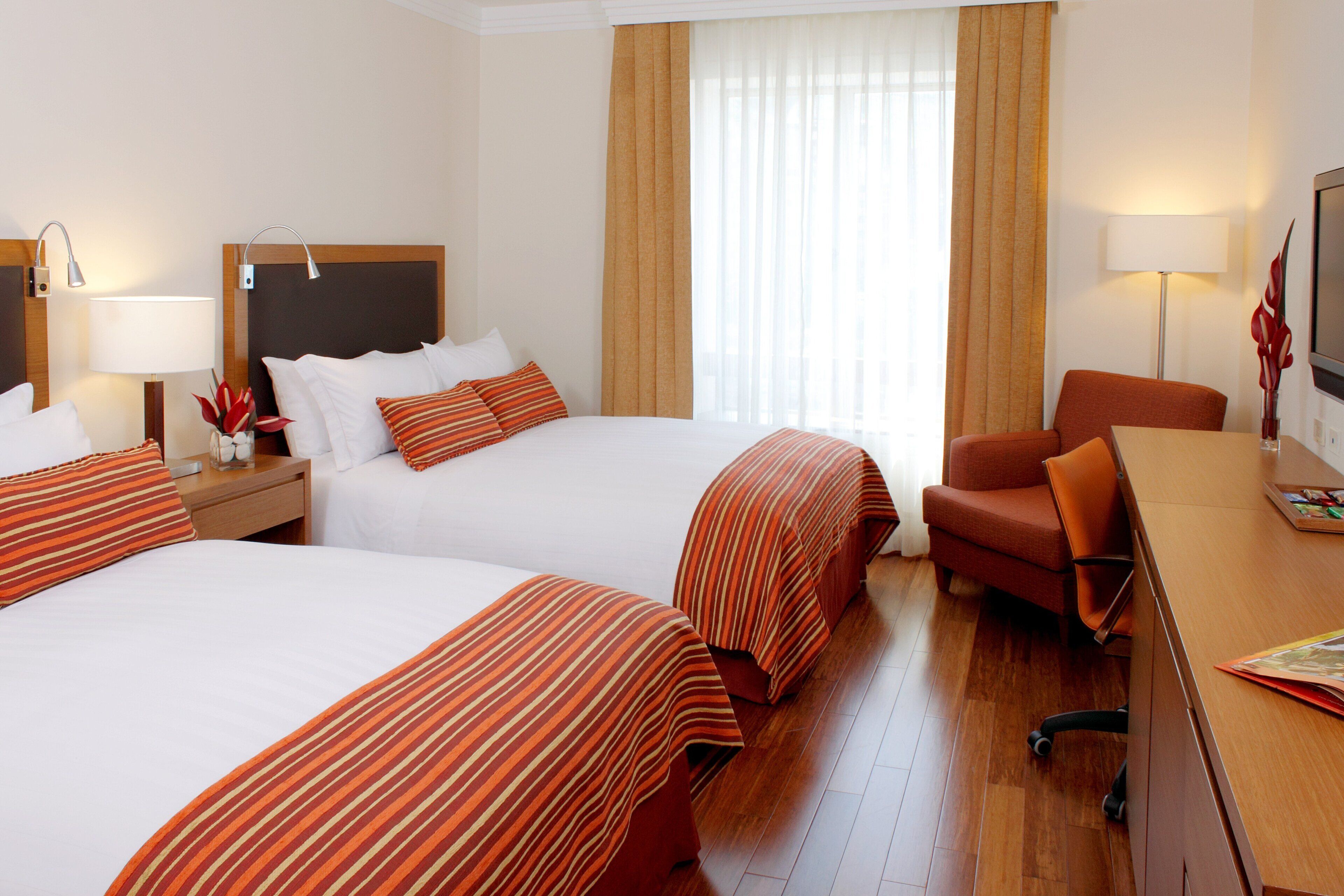 Ein Hotelzimmer mit zwei Betten, einem Schreibtisch, einem Stuhl, einem Fernseher und einem Fenster mit Vorhängen.