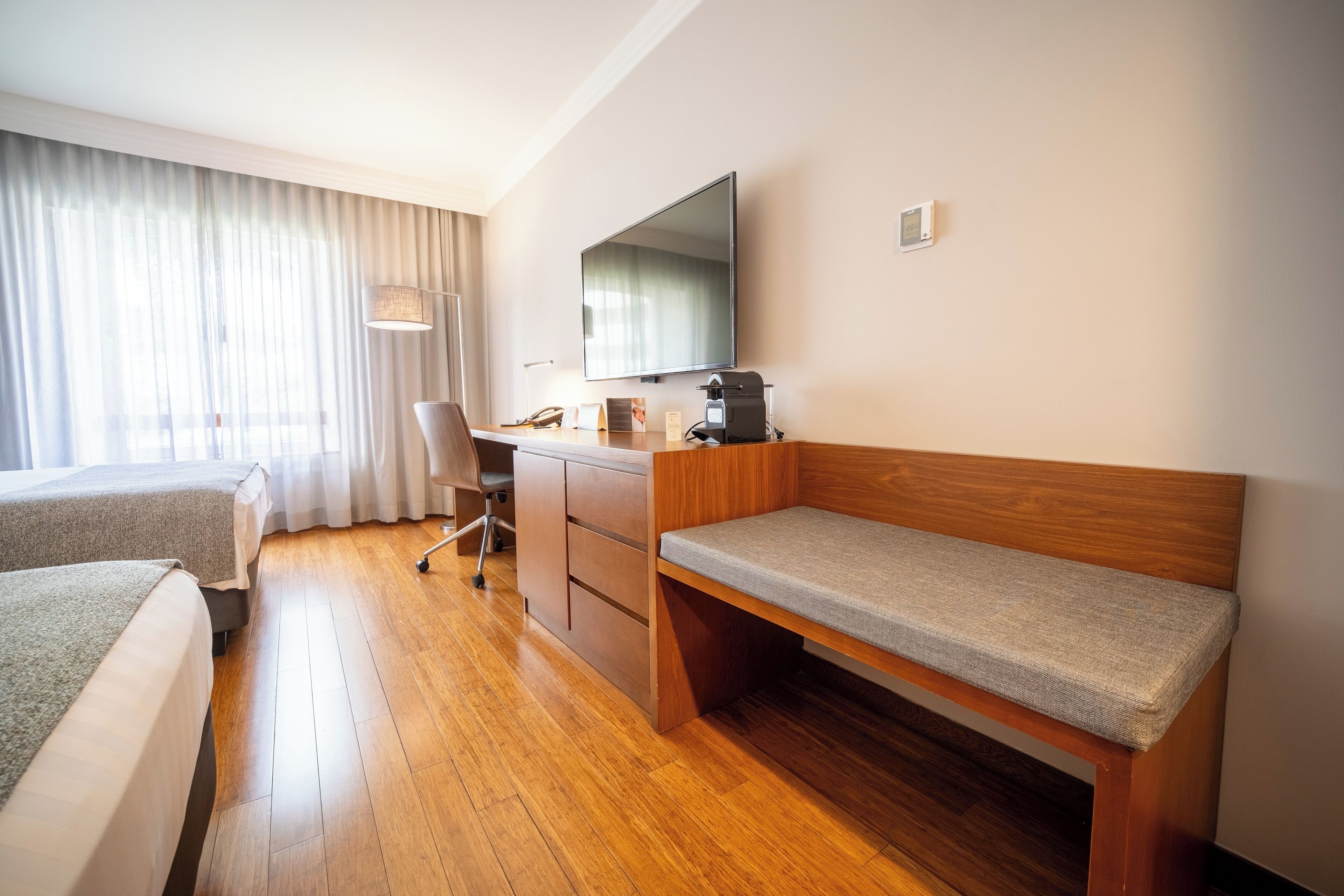 Habitación de hotel con un escritorio de madera, una cama, una silla, televisión y una ventana con cortinas.