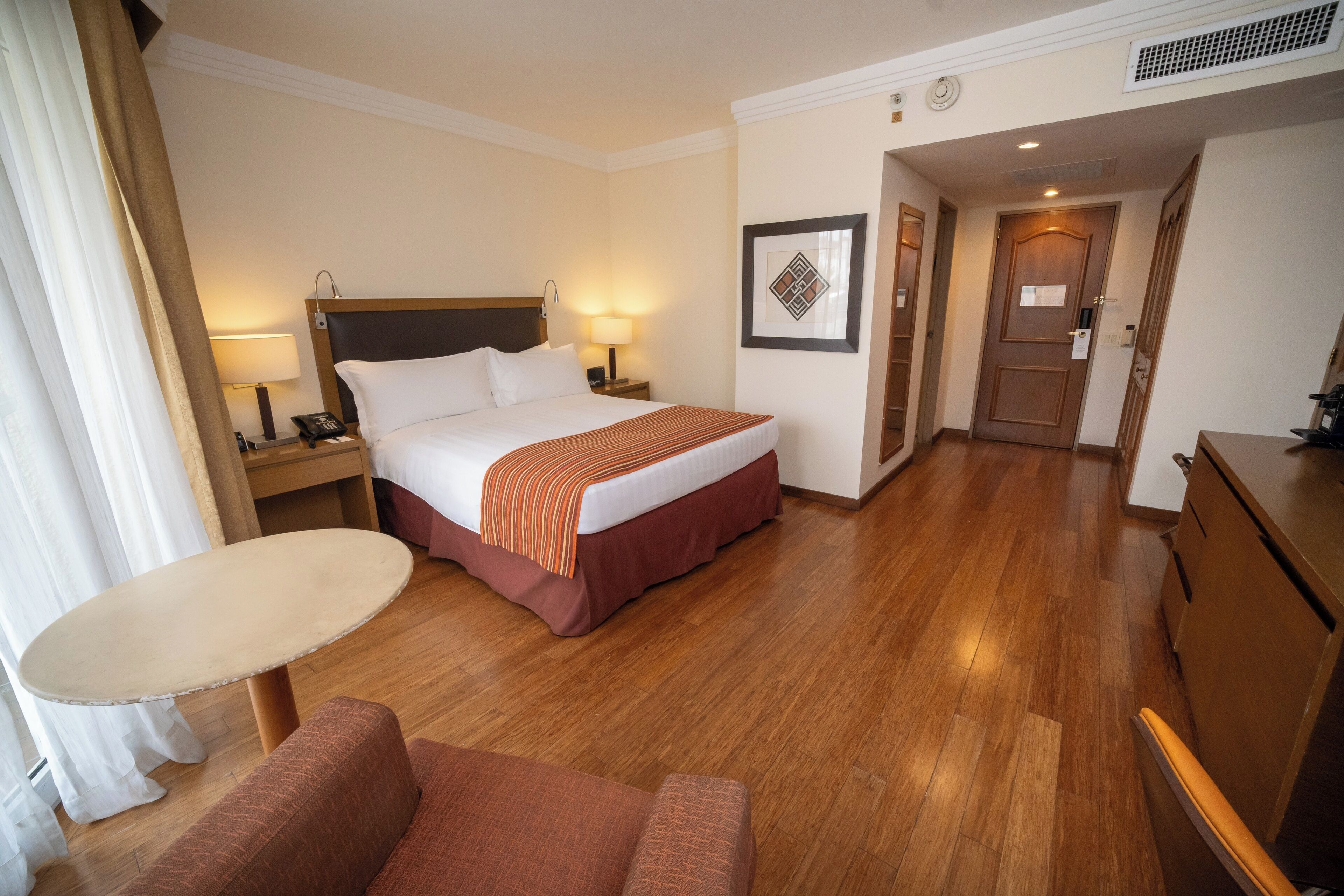 A hotel room with a bed, bedside table, lamp, chair, and a small table.