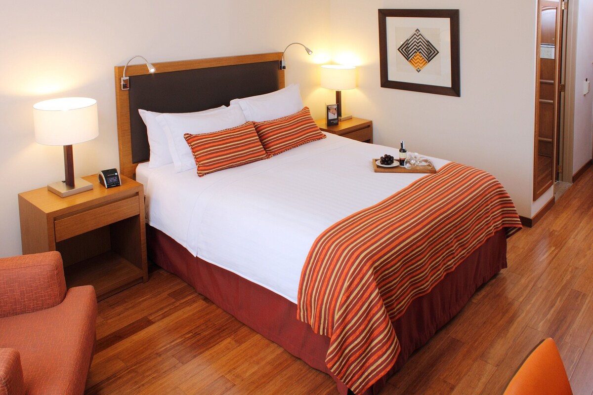 Une chambre d’hôtel avec un lit, des tables de chevet, une lampe, une chaise et un tableau encadré au mur.