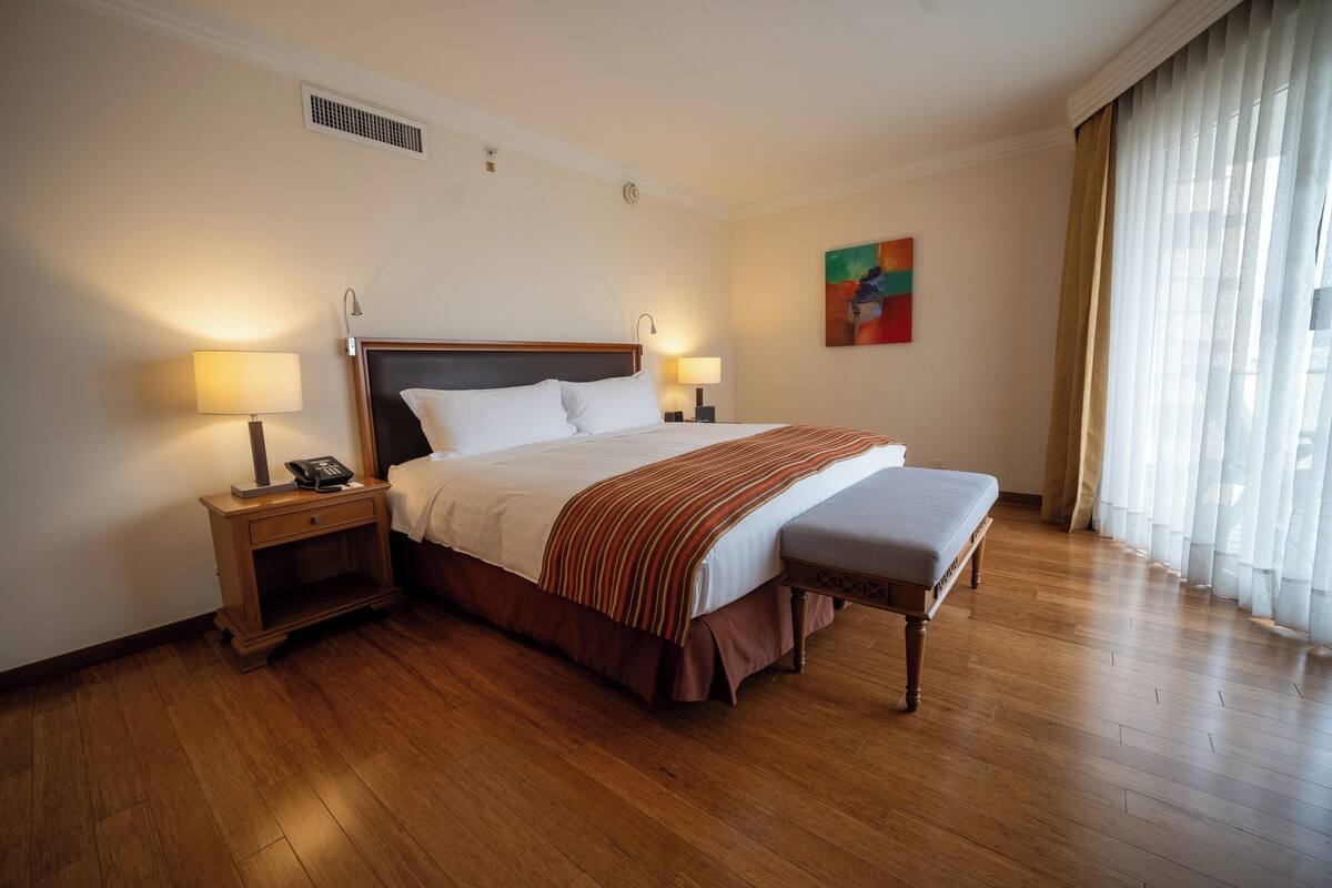 A hotel room with a large bed, two bedside tables with lamps, a bench, a painting on the wall, and a window with curtains.