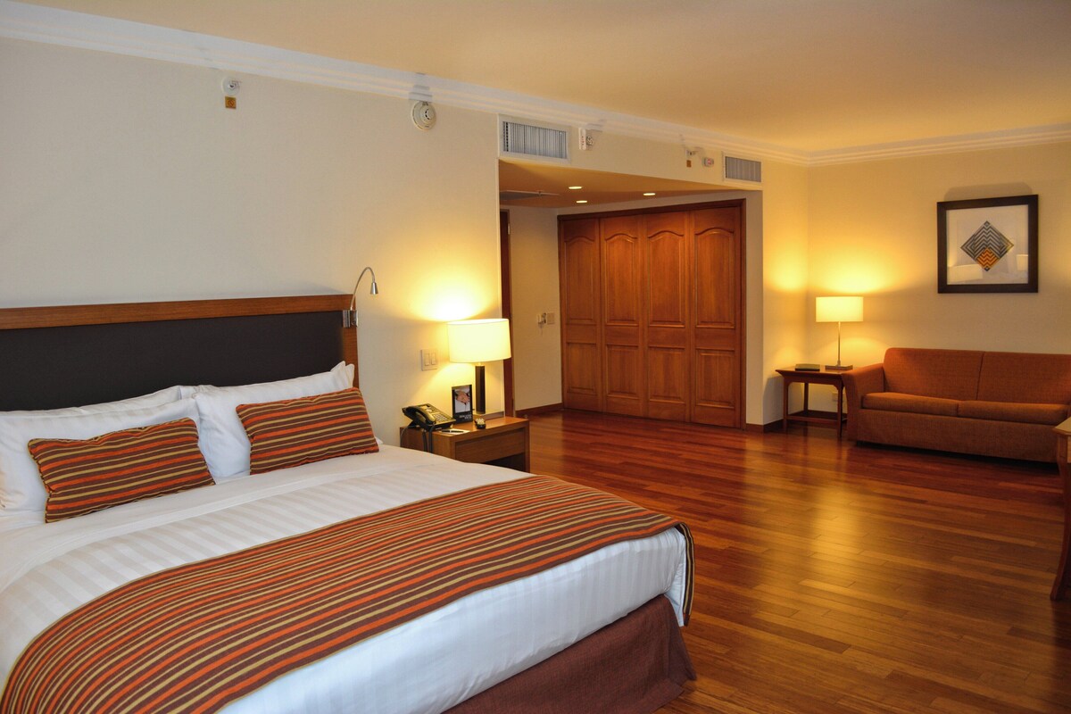 Une chambre d’hôtel avec un grand lit, une armoire en bois, un canapé, une table de chevet avec une lampe et un tableau encadré au mur.